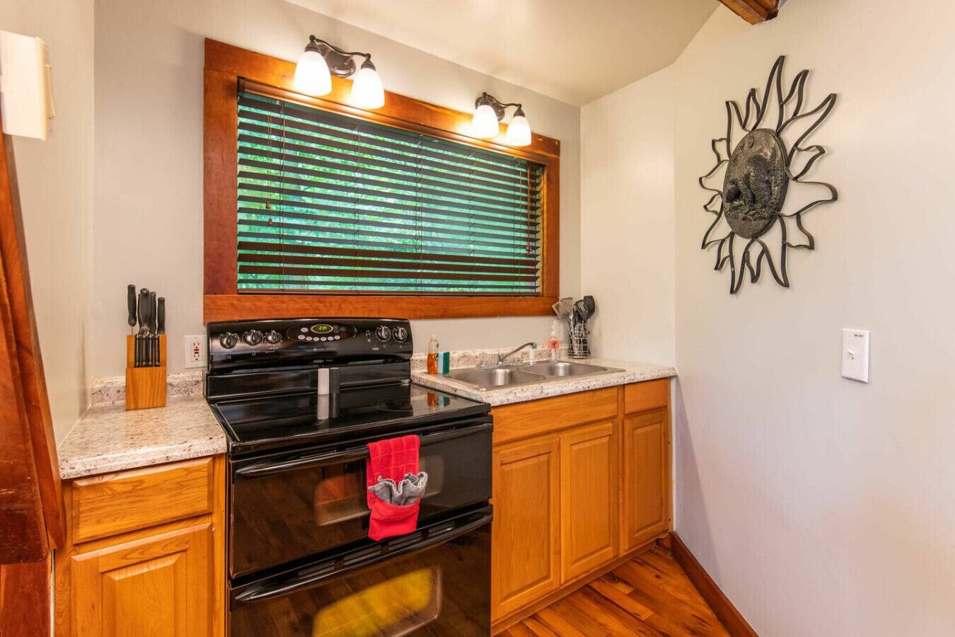 Una cocina con armarios de madera, estufa negra, ventana con persianas, pileta y un adorno de pared.