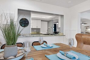 Un comedor con una mesa de madera dispuesta para dos comensales, un florero con conchas marinas y un espejo que refleja la cocina.