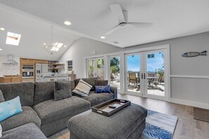 Ein modernes Wohnzimmer mit einem grauen Ecksofa, einem Couchtisch und einem Deckenventilator. Eine Glastür öffnet sich zu einer Außenpatiofläche.