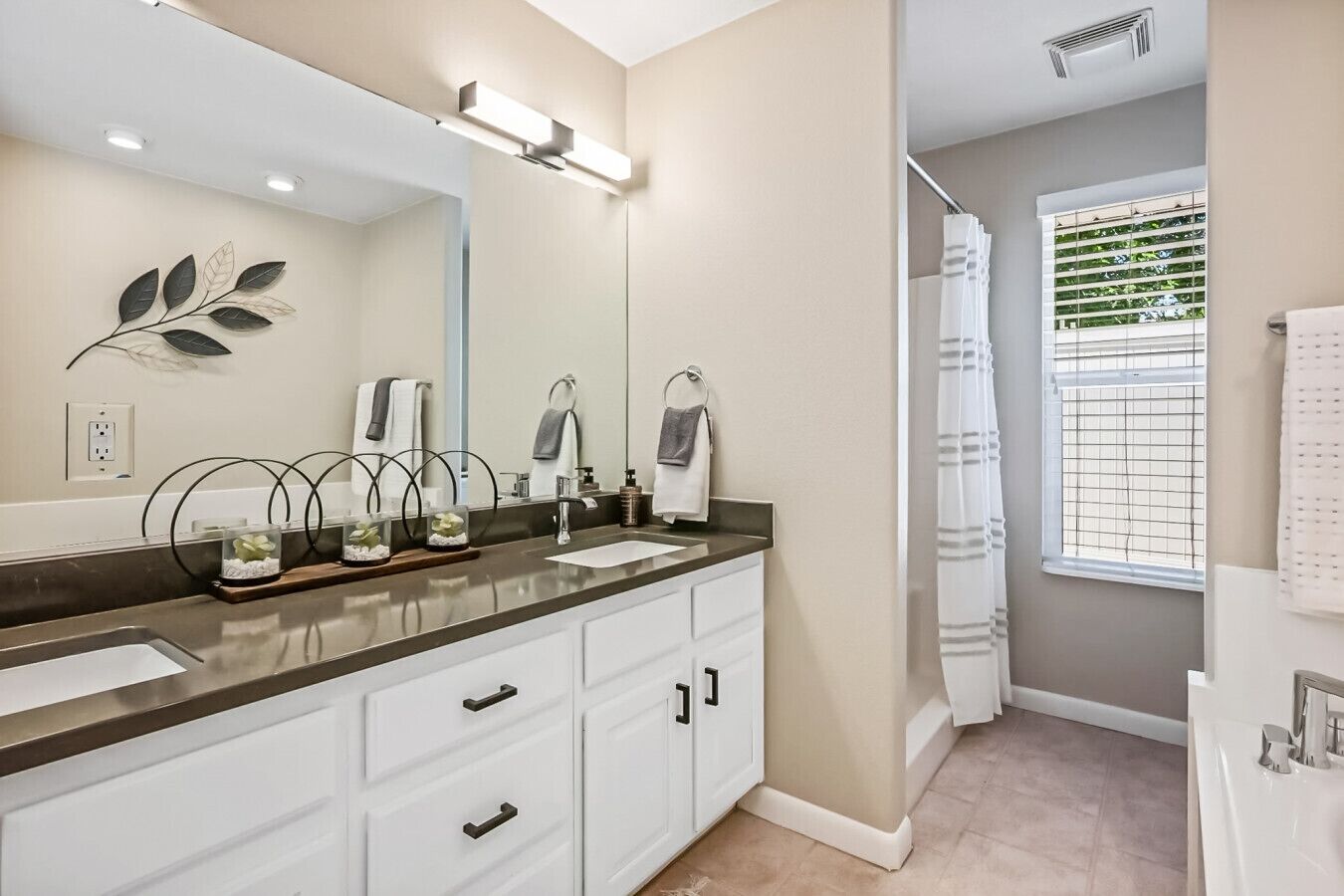 A modern bathroom with a large mirror, two sinks, a shower, and a window with blinds.