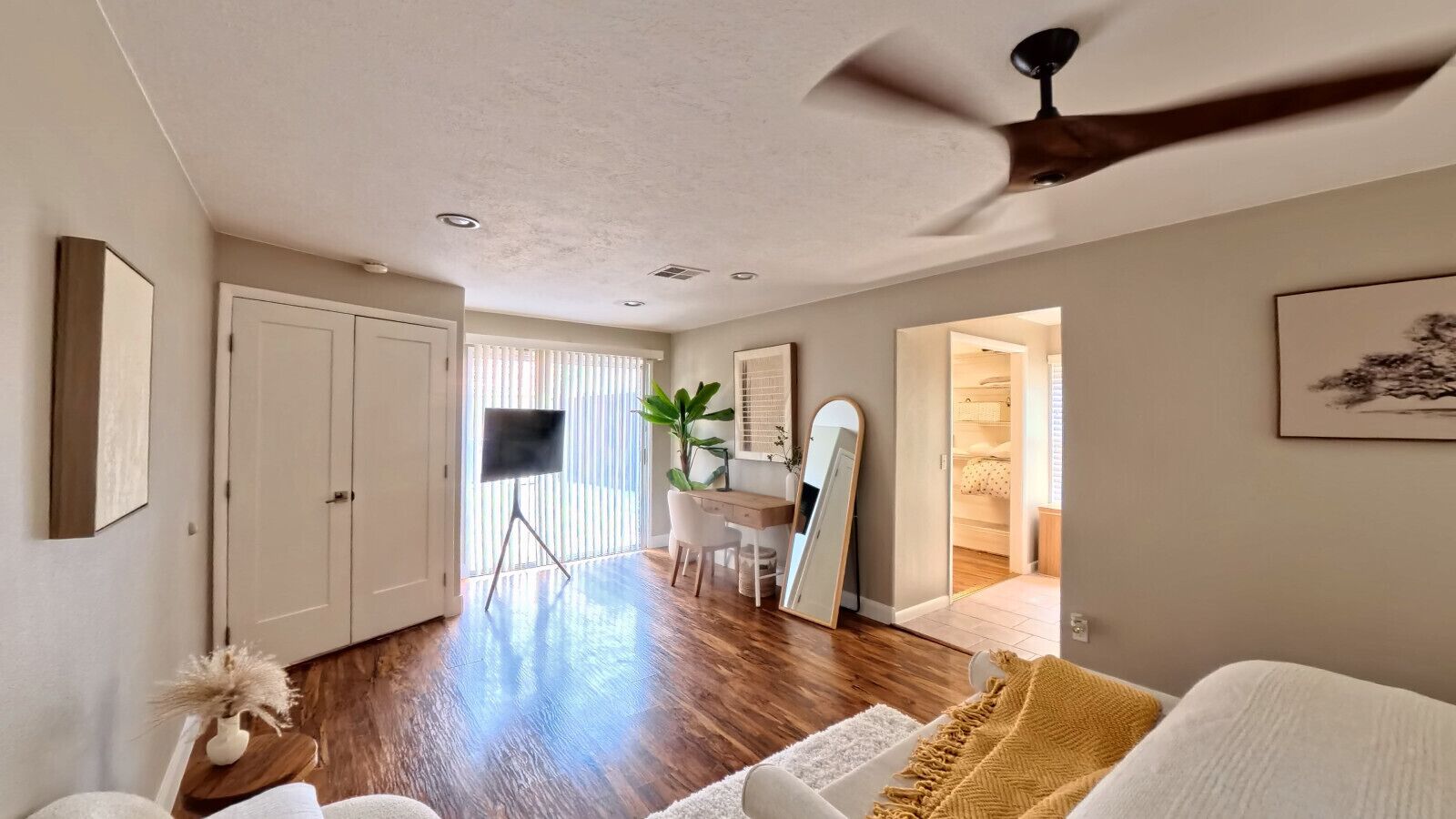 A modern living room with hardwood flooring, a ceiling fan, a desk with a chair, a large mirror, and a door leading to another room.