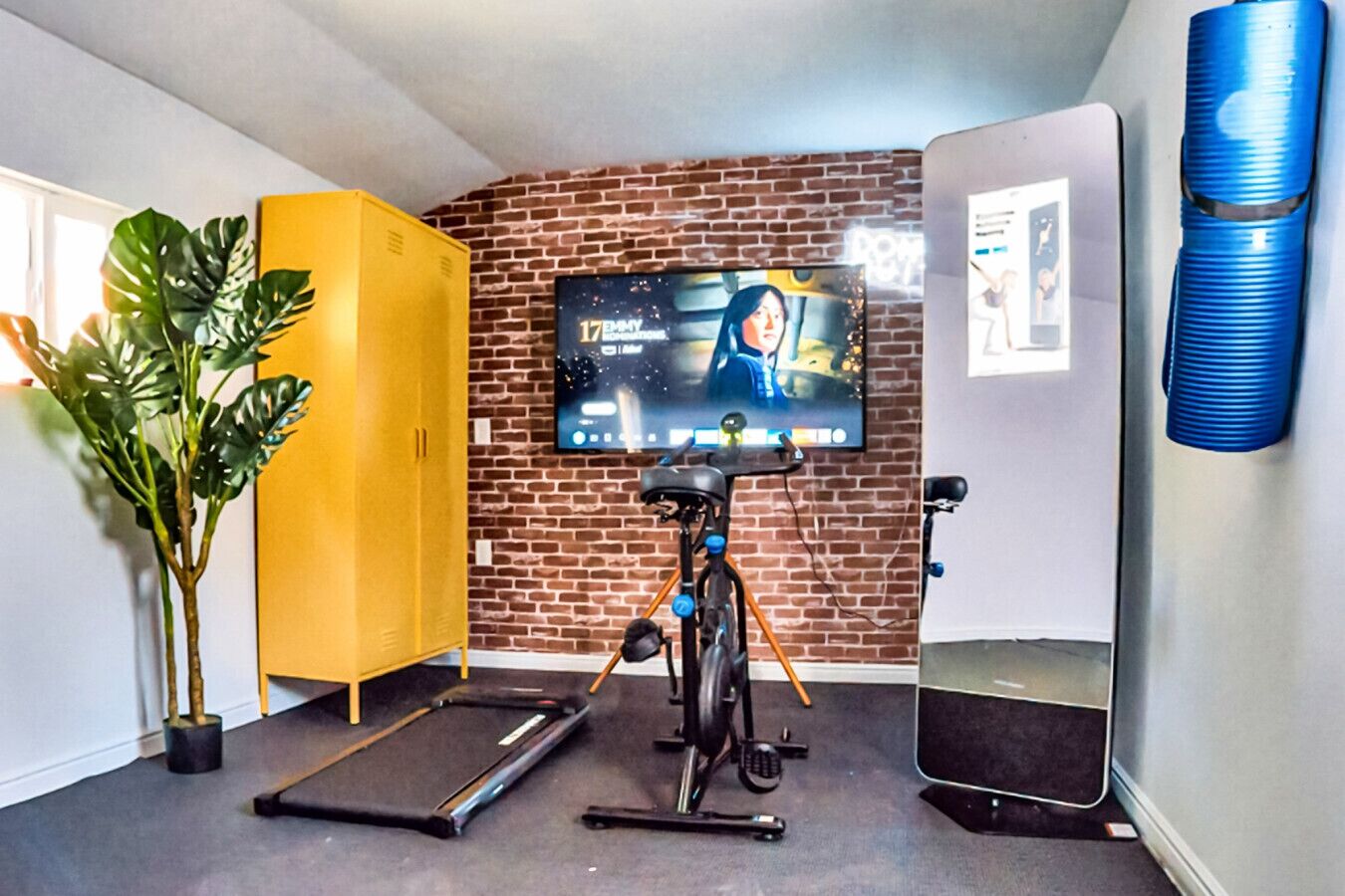 A room with a brick wall, a TV mounted on it, a treadmill, a stationary bike, a yellow cabinet, and a potted plant.