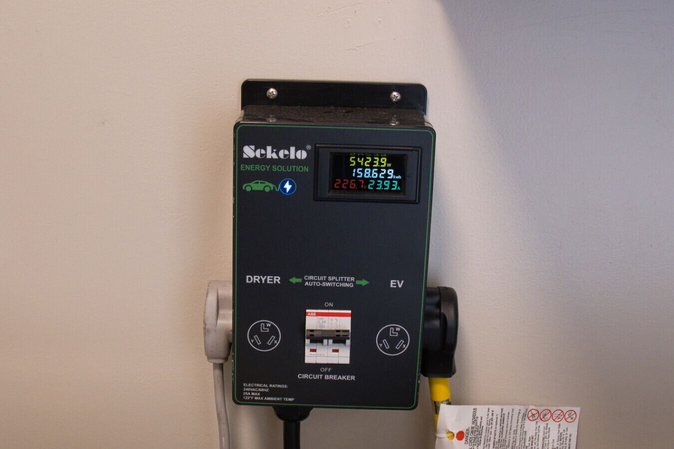 Un medidor de energía Sekel con pantalla que muestra el consumo energético en kWh, conectado a una secadora y un cargador para vehículos eléctricos.