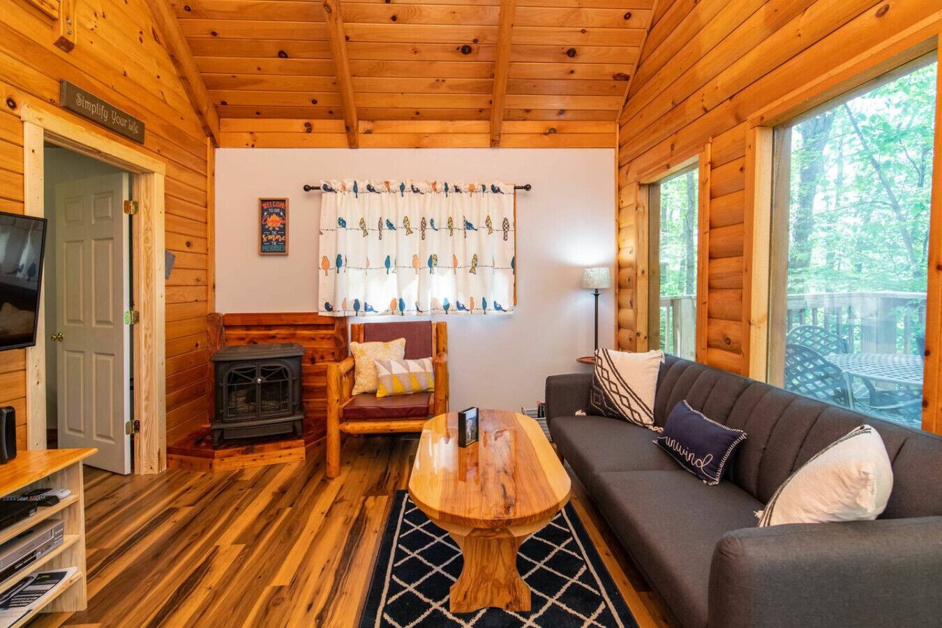 A cozy cabin interior with a wooden floor, a sofa, a chair, a small table, a fireplace, and a TV.