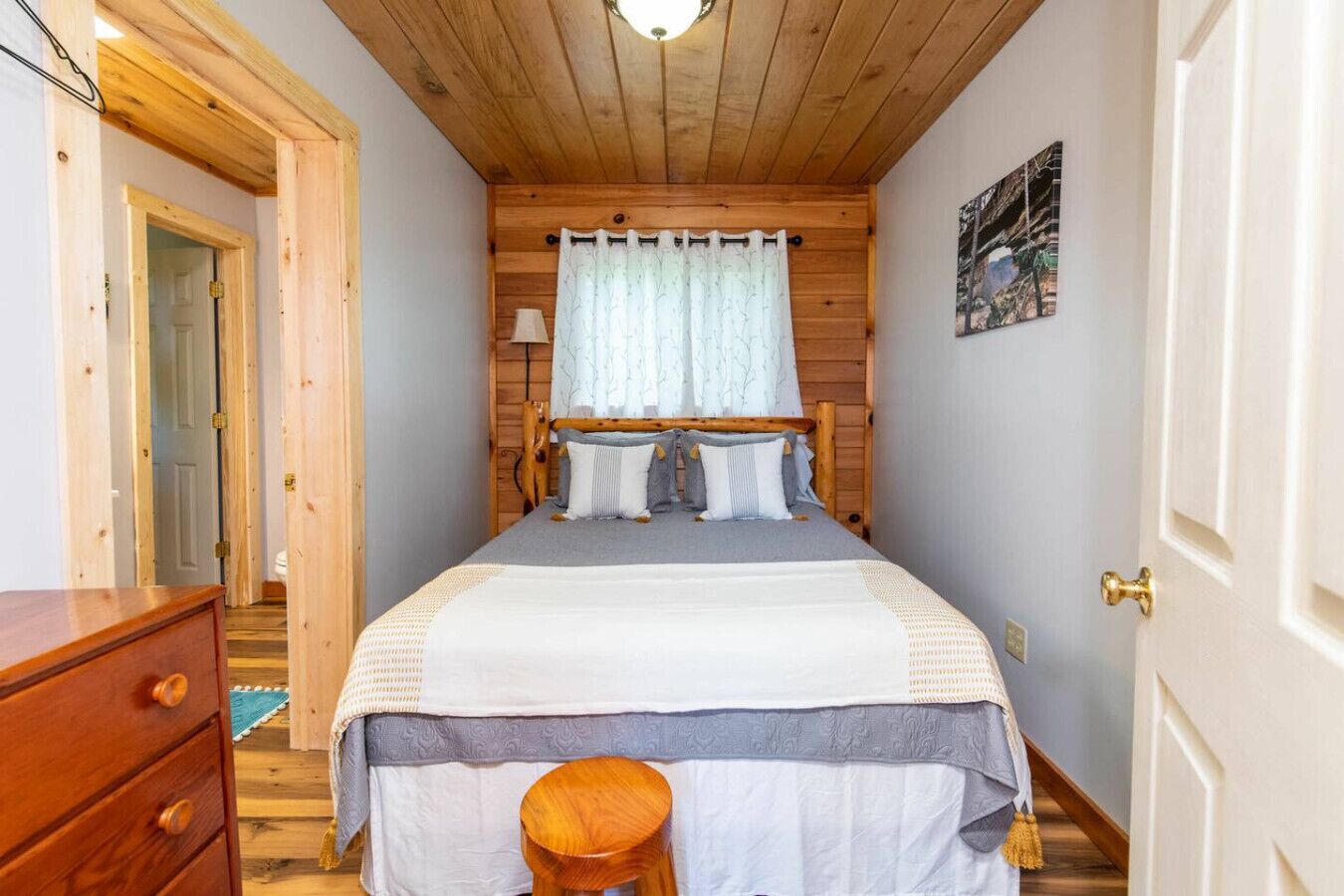 A bedroom with a wooden ceiling, a bed with a white and gray bedspread, a wooden nightstand, and a wooden stool.