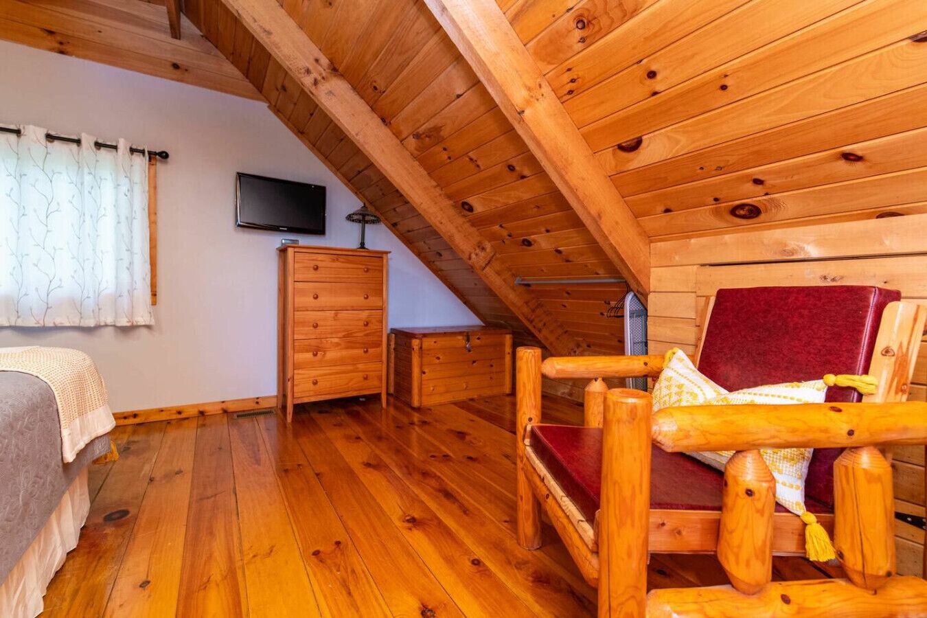 A cozy room with wooden floors, a red cushioned chair, a wooden dresser, and a television on the wall.