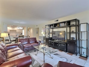 A living room with a leather sofa, a glass coffee table, and a TV on a black entertainment unit. - Bay Oaks - E53 (Siesta Key)