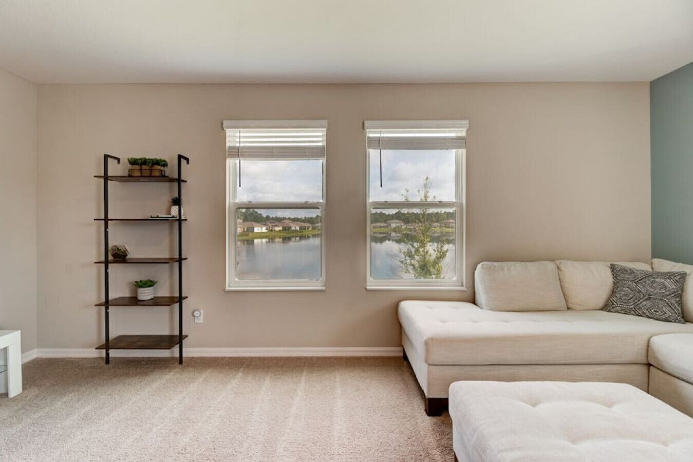 Una sala de estar con un sofá beige, un otoman blanco, una estantería y dos ventanas con vista al lago.