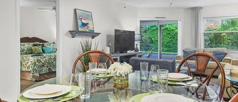 A dining area with a glass table set for four, surrounded by wooden chairs. A ceiling fan and a television are visible in the background.