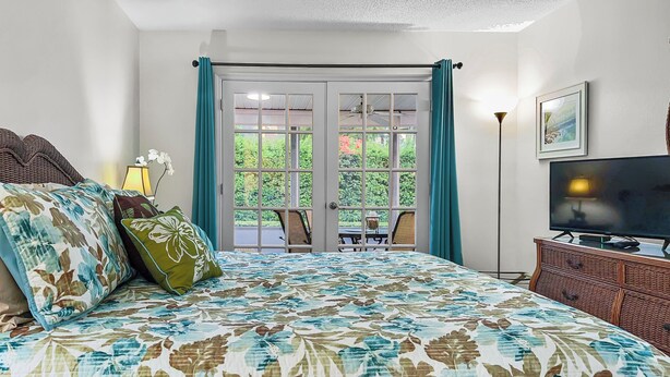 A bedroom with a large bed, a television, and a view of a garden through a glass door. - Bayside Pointe 3 - 1302 (Siesta Key)
