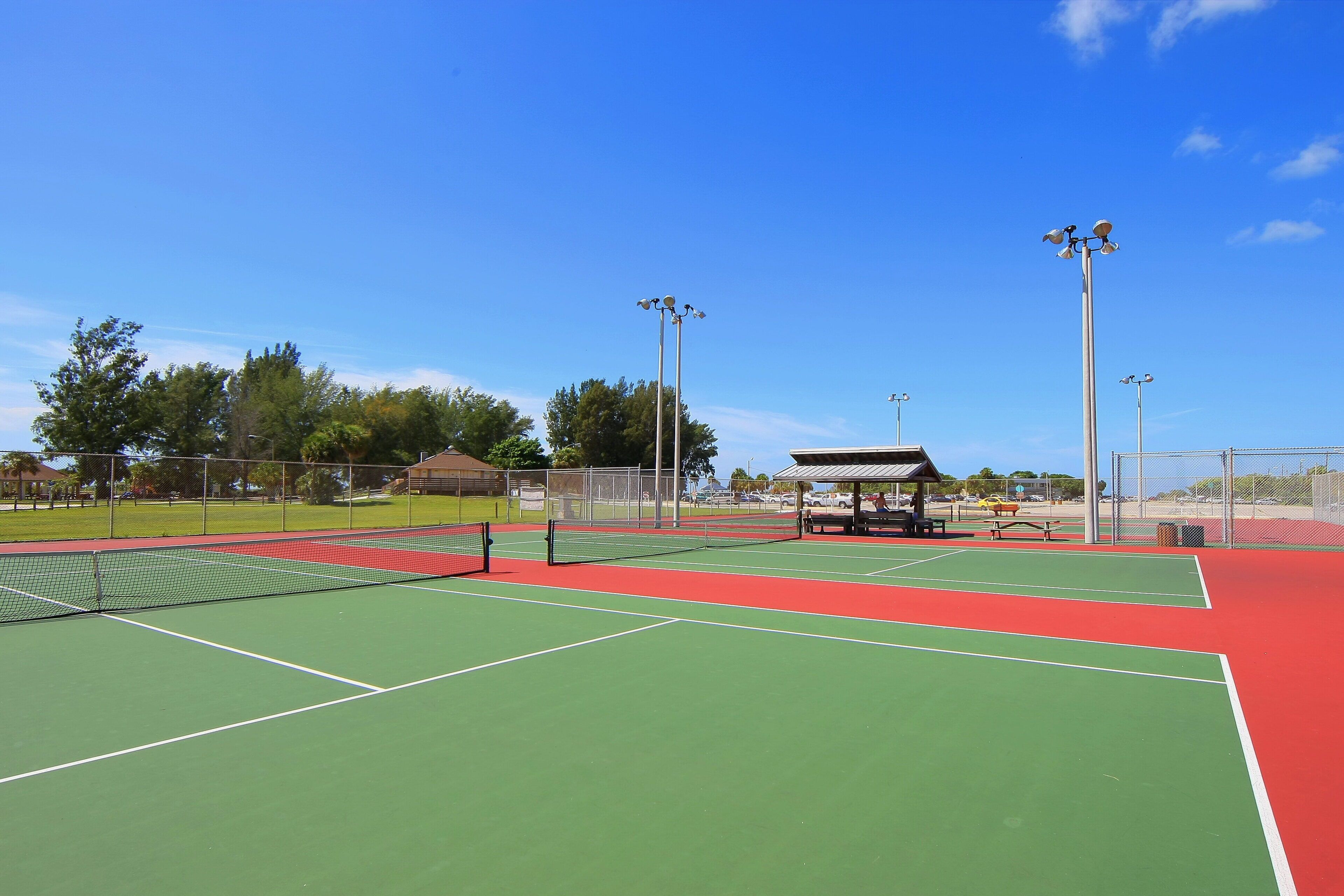 A tennis court with a net, surrounded by a fence, and a pavilion in the background.