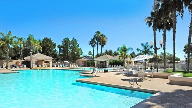 A swimming pool with lounge chairs and umbrellas, surrounded by palm trees and covered structures. - Michigan Sun Lakes (Chandler)