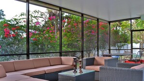 A covered outdoor seating area with a sofa, chairs, and tables, overlooking a garden with pink flowers. - Edgemere (Siesta Key)