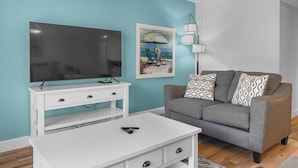 A living room with a flat-screen TV, a white TV stand, a grey sofa with patterned cushions, and a floor lamp. - Midnight Cove II - Sunrise (Siesta Key)