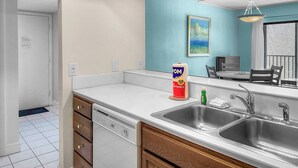 A modern kitchen with a dishwasher, two sinks, and a dining area with a chair and table. - Midnight Cove II - Sunrise (Siesta Key)