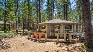 A wooden gazebo with a glass enclosure, surrounded by a deck with orange chairs and a table, set in a forested area. - Carousel House (Bend)