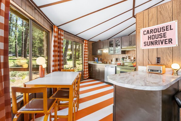 A modern kitchen with wooden cabinets, a dining area with striped flooring, and a sign reading "CAROUSEL HOUSE SUNRIVER". - Carousel House (Bend)