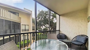 A balcony with a glass table and two chairs, a patterned rug, and a view of neighboring buildings and trees. - Bay Oaks - E82 (Siesta Key)