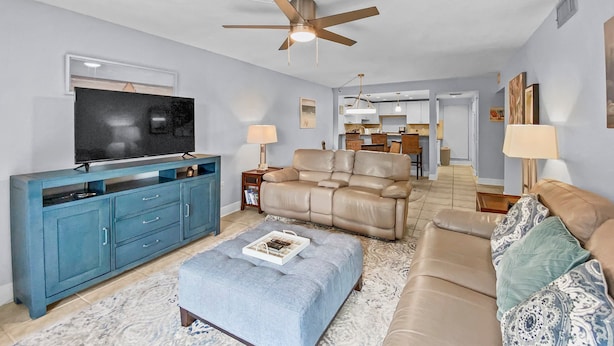 A living room with a TV, blue entertainment unit, beige sofa, ottoman, and a ceiling fan. - Bay Oaks - E82 (Siesta Key)