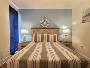 Une chambre avec une couvre-lit à rayures, une tête de lit en bois, deux lampes de chevet et une décoration représentant un gouvernail de bateau accrochée au mur.