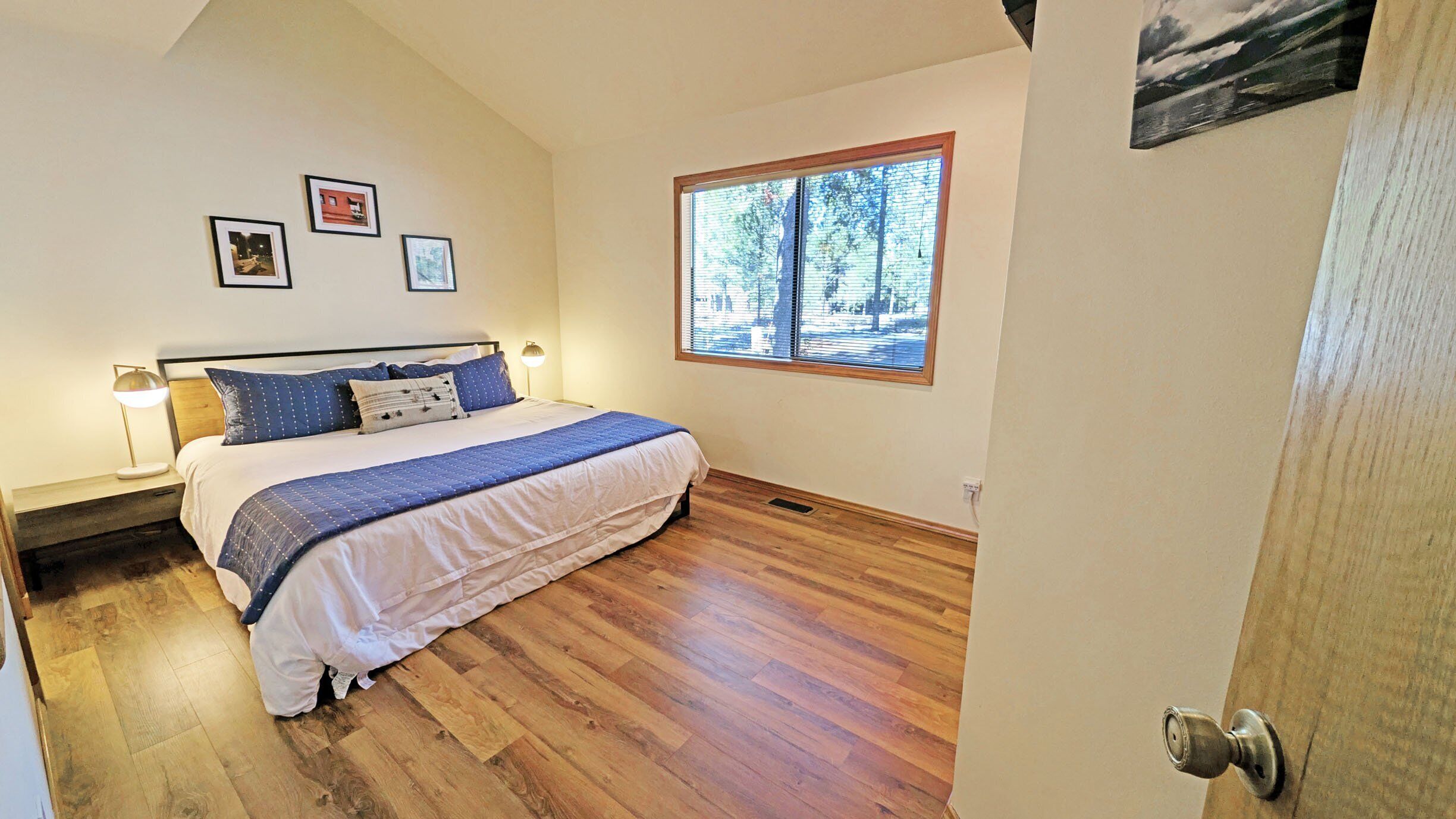 A bedroom with a bed, wooden flooring, a window with blinds, and a door.