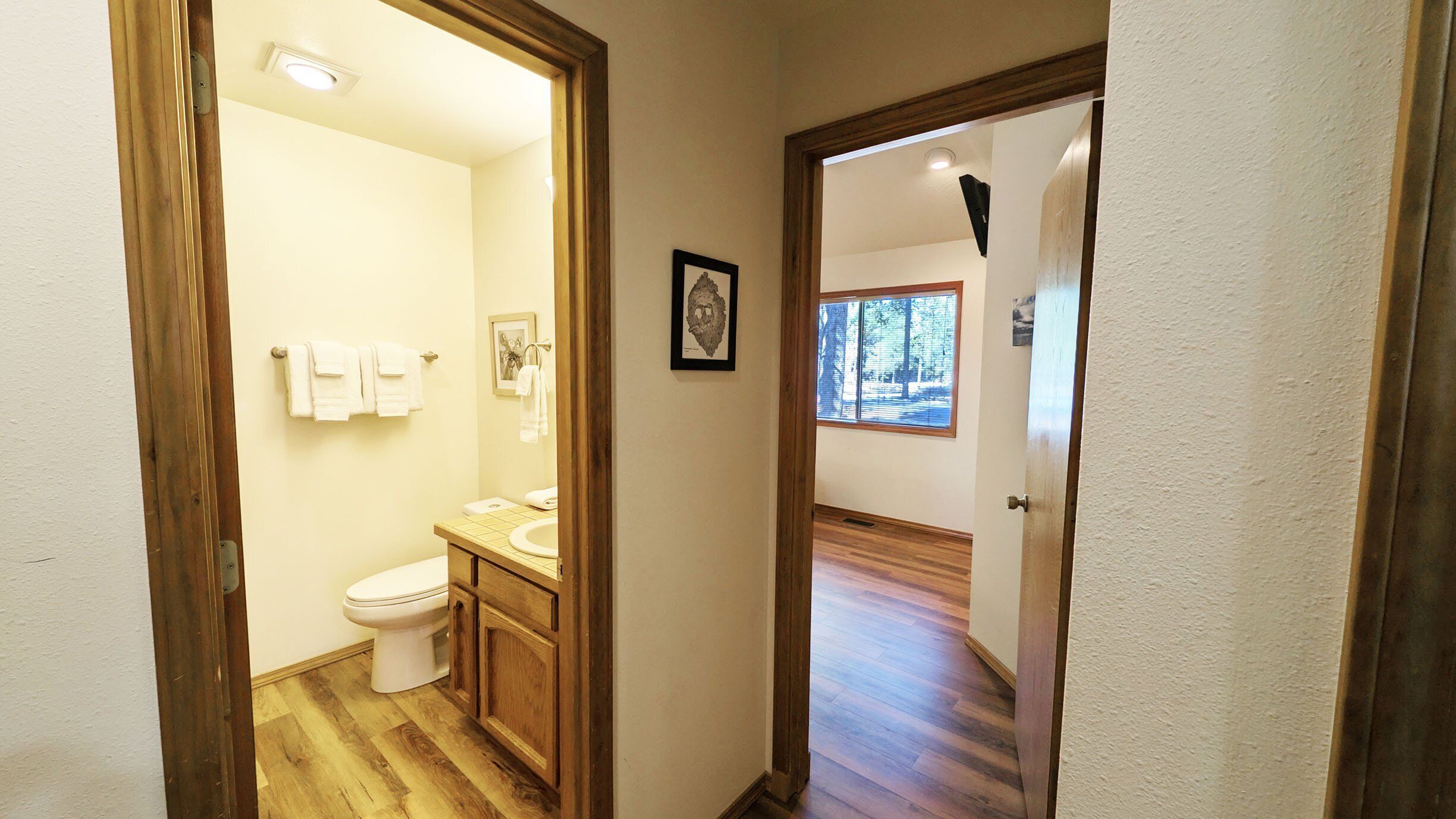 Une salle de bain avec des toilettes, un lavabo et des meubles en bois. Un tableau encadré est accroché au mur, et une fenêtre offre une vue sur les arbres.