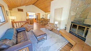 Amplia sala de estar con chimenea de piedra, sofá de cuero, mesa de comedor de madera y cocina al fondo.