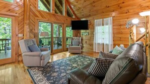 A wooden cabin interior with a living room, a television, and a deck with outdoor seating. - Hot Tub & Wifi - Birdsong - Red River Gorge, KY (Stanton)