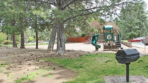 Une aire de jeux avec un toboggan vert et une structure de jeu, entourée d’arbres et d’une pelouse.