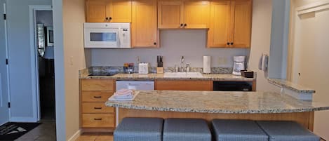 A compact kitchen with wooden cabinets, a microwave, a coffee maker, and a built-in dishwasher. There are three bar stools and a small countertop.