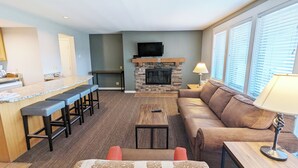 A modern living room with a stone fireplace, a bar area with stools, a sofa, and a TV mounted above the fireplace. - 7th Mtn 1535f (Bend)