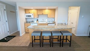 A modern kitchen with granite countertops, wooden cabinets, and a built-in microwave. There are three bar stools with cushioned seats. - 7th Mtn 1535f (Bend)