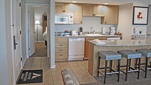 A modern kitchen with wooden cabinets, a built-in microwave, a dishwasher, and a granite countertop. There are three bar stools and a bench at the island. - 7th Mtn 1235a (Bend)