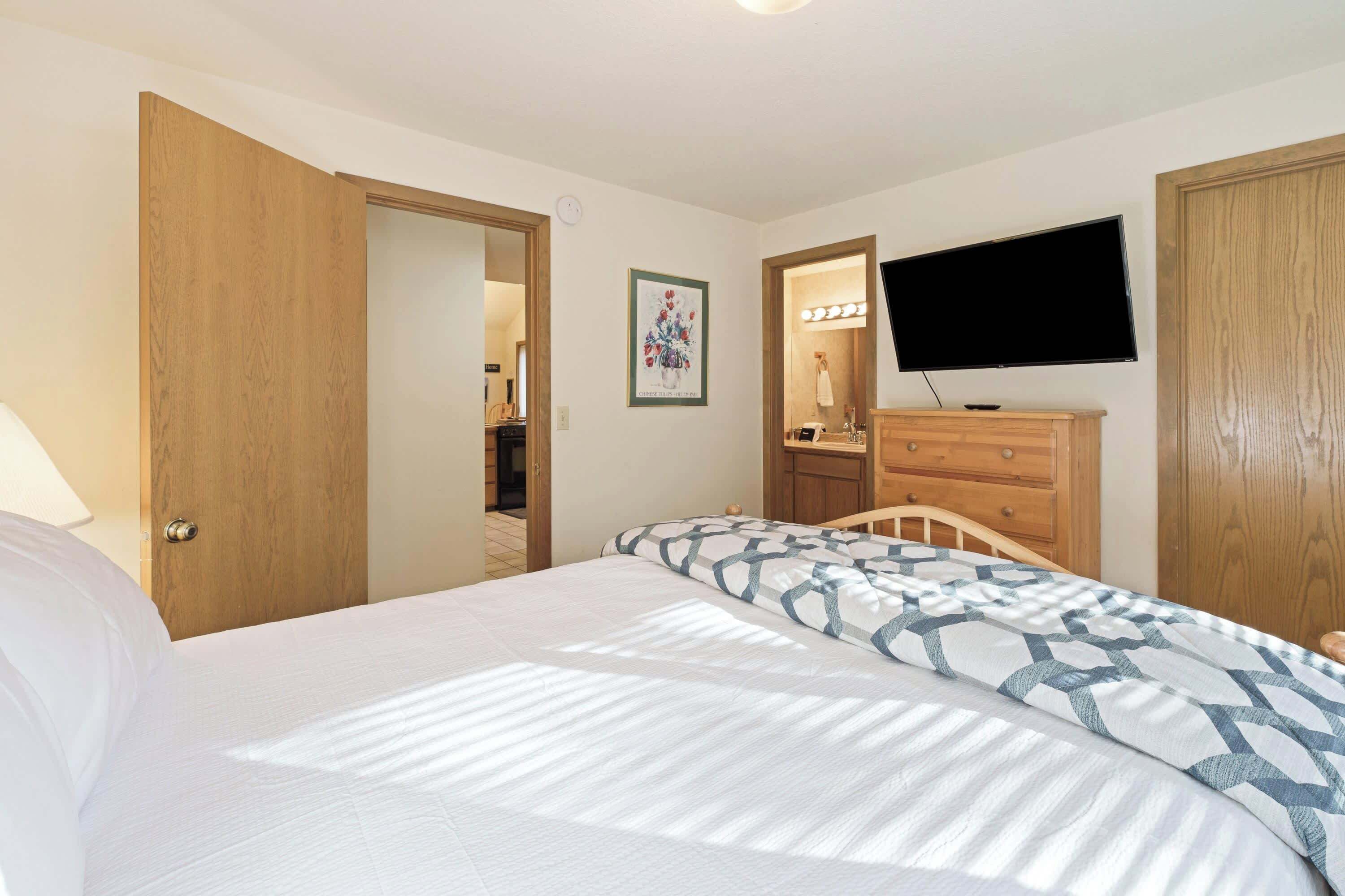 A bedroom with a bed, a television mounted on the wall, a dresser, and a door leading to another room.
