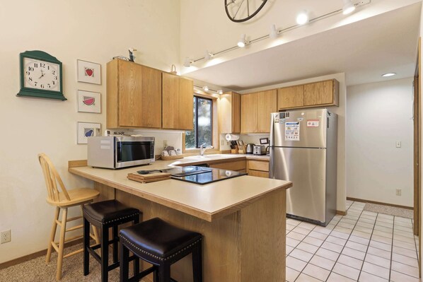 A modern kitchen with wooden cabinets, a stainless steel refrigerator, a microwave, and a sink. There are two stools and a clock on the wall. - Blue Goose With Hot Tub & Sharc Access (Bend)