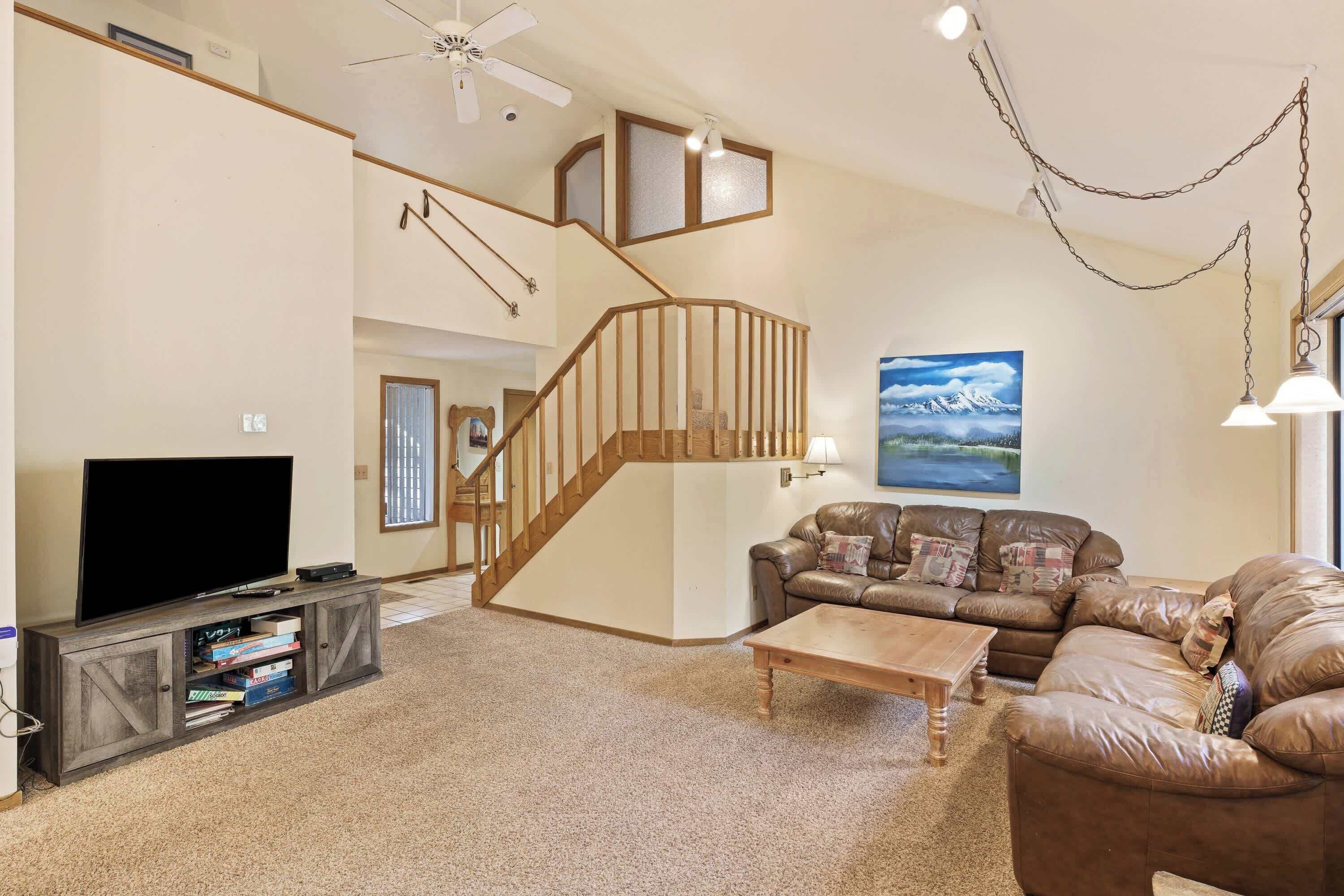 A spacious living room with a wooden staircase, a leather sofa, a flat-screen TV, and a bookshelf.
