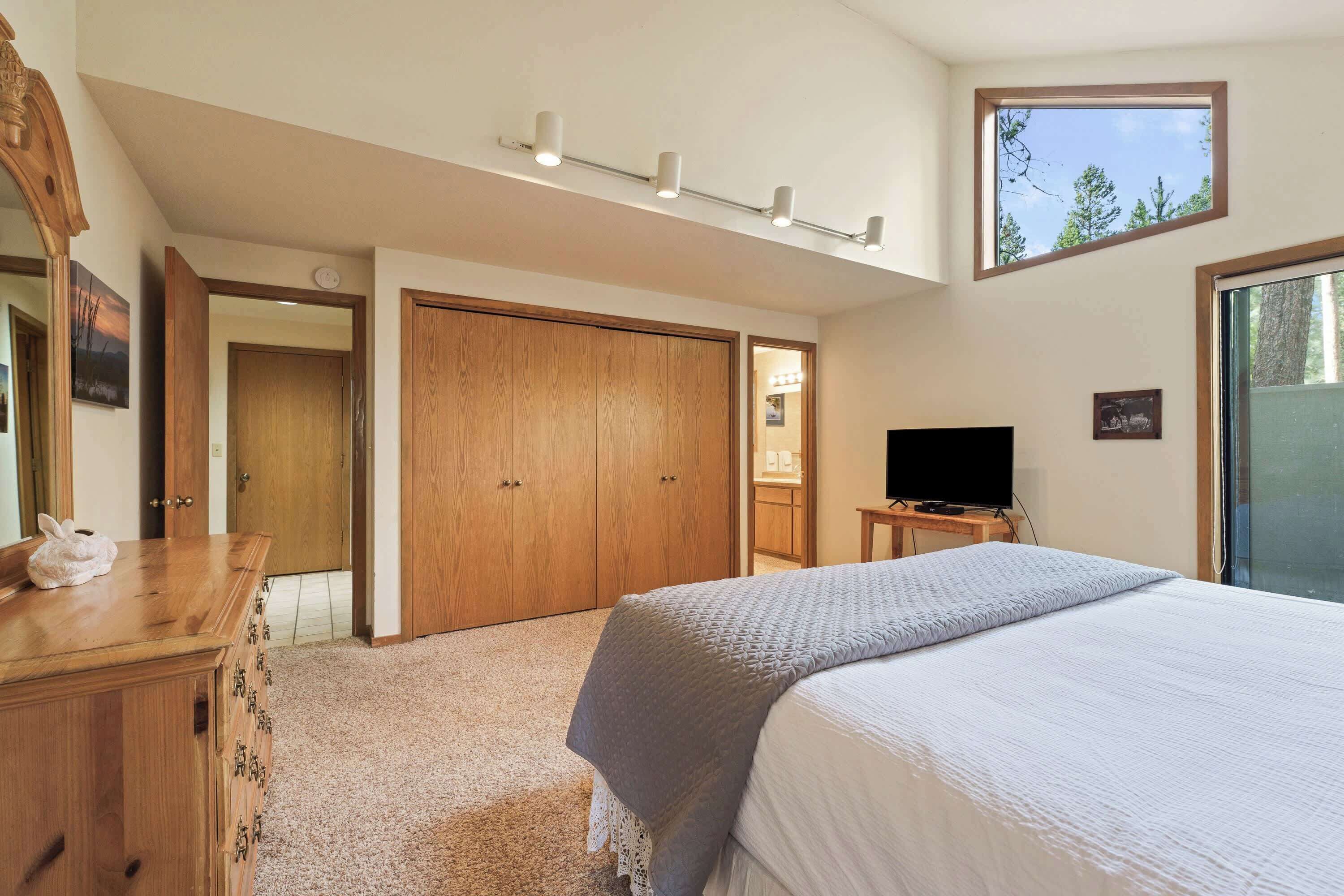 A bedroom with a bed, a wooden dresser, a TV, and a large window.