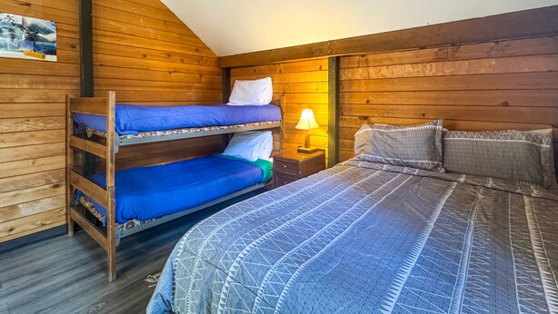 Ein Zimmer mit Holzwänden, einem Etagenbett, einem Einzelbett und einem Nachttisch mit Lampe.