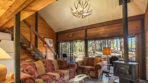 Ein gemütliches Blockhaus-Interieur mit einer Couch, einem Holztisch, einem Kamin und einer Treppe zu einer Terrasse mit Blick auf den Wald.