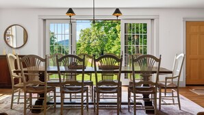 A dining area with a table and chairs, large windows with a view of greenery, and a door. - 52-acre Berkshires Farmhouse Fire Pit, Outdoor Dining + Trails Copper Kettle Farm by Avantstay (Ashley Falls)