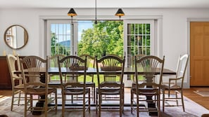 A dining area with a table and chairs, large windows with a view of greenery, and a door. - 52-acre Berkshires Farmhouse Fire Pit, Outdoor Dining + Trails Copper Kettle Farm by Avantstay (Ashley Falls)
