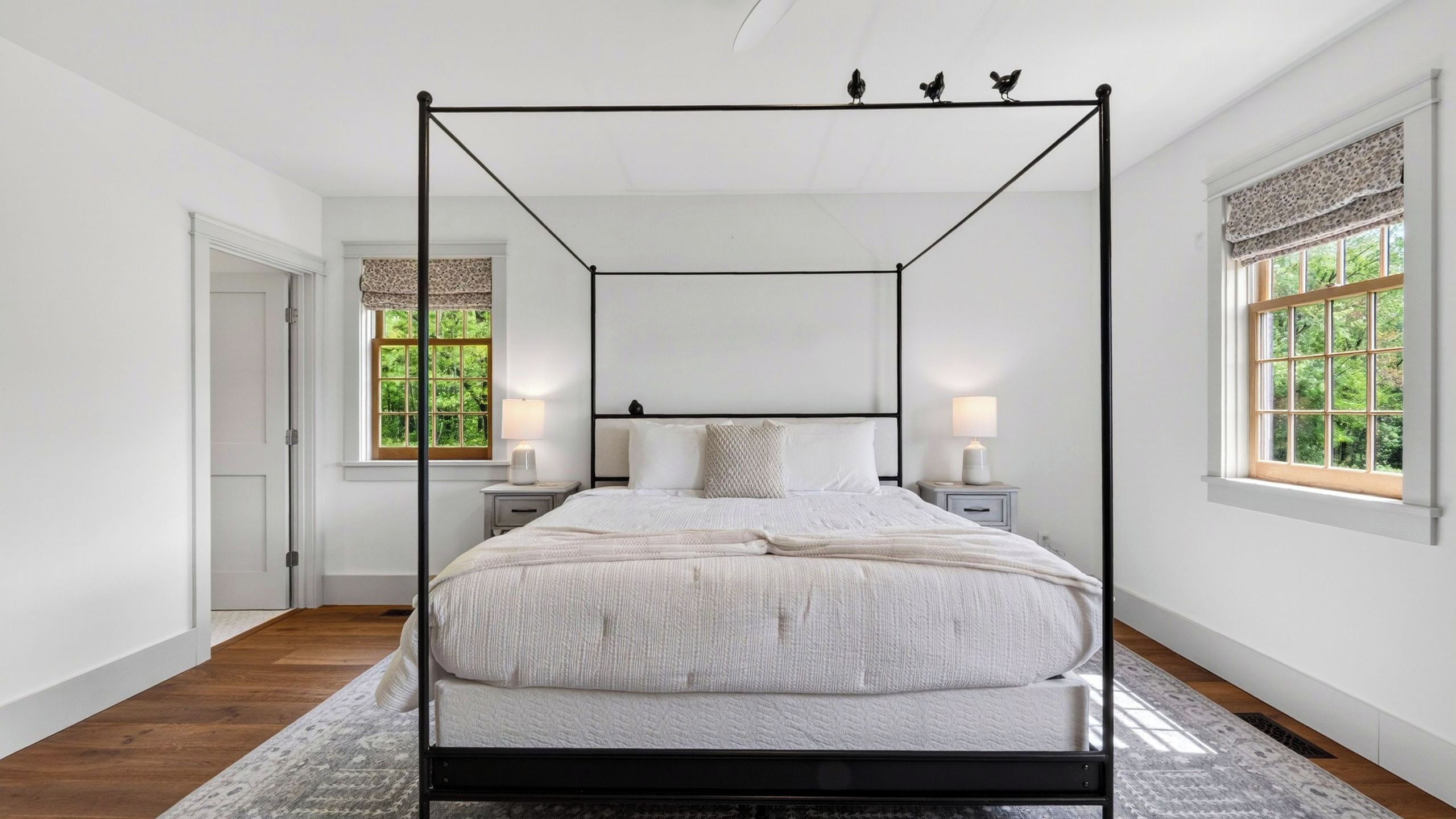 A four-poster bed in a room with two bedside tables, a door, and windows with curtains.