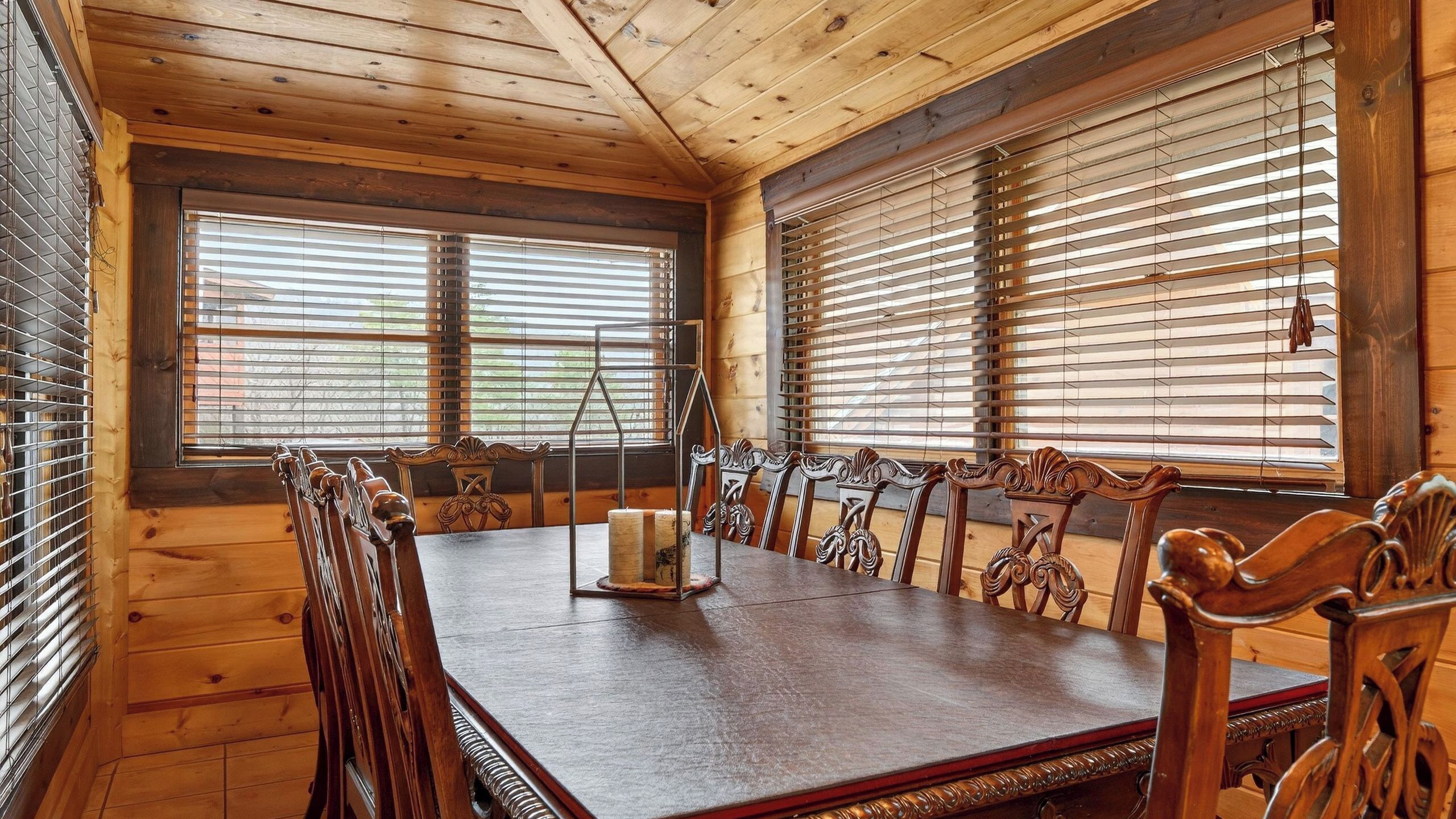 A wooden-paneled room with a large dining table and chairs, large windows with blinds, and a candle holder on the table.