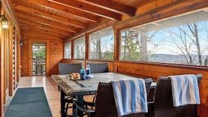 A wooden cabin with a dining table, chairs, and a view of trees and mountains.