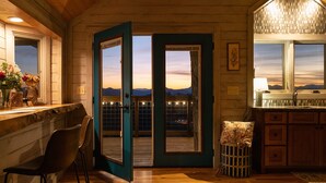 A cozy cabin interior with a wooden floor, a bar area, a dining space with chairs, and a large door leading to a balcony with a view of the mountains. - Take in the rustic, fairy tale vibe of this custom-made treehouse in the midst of towering white oaks. Recline in a rocking chair and watch the sunset over the mountains, or try your hand at some stargazing from the comfort of the deluxe double bed.< (Asheville)