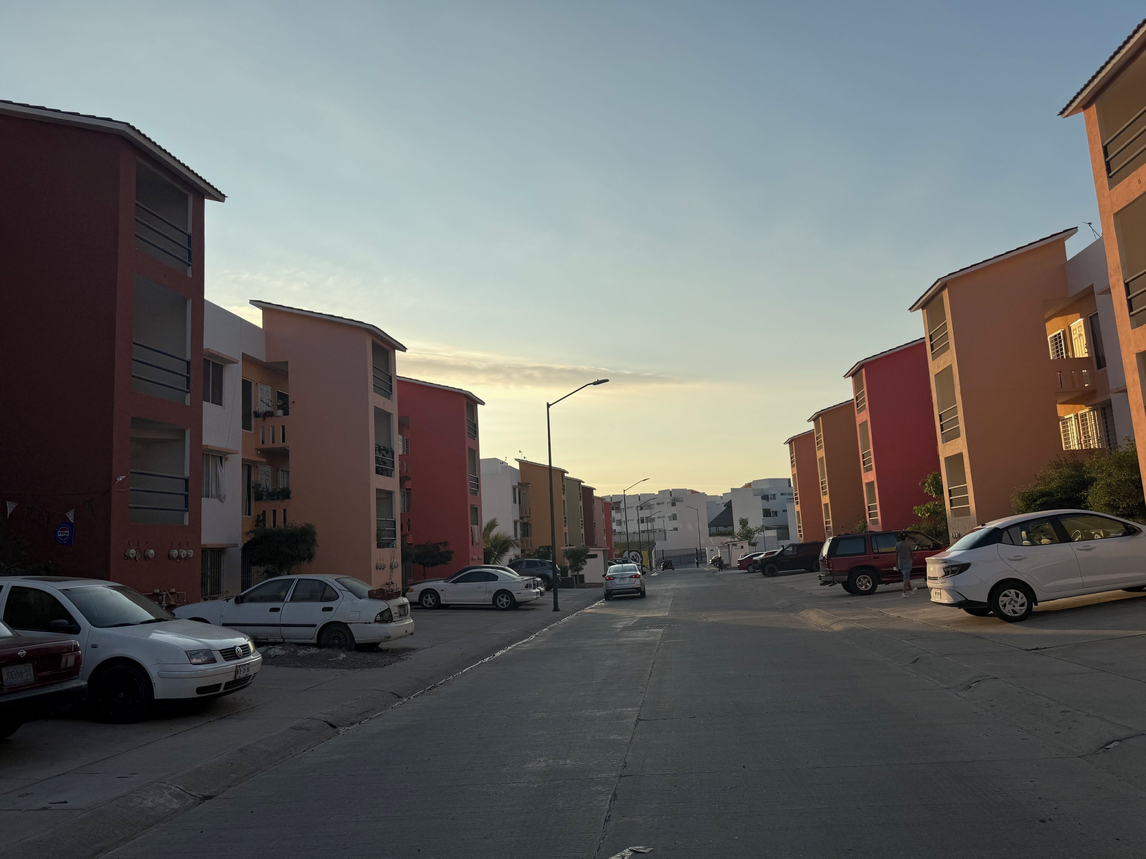 Una calle residencial con edificios de varios pisos, coches aparcados y cielo despejado.