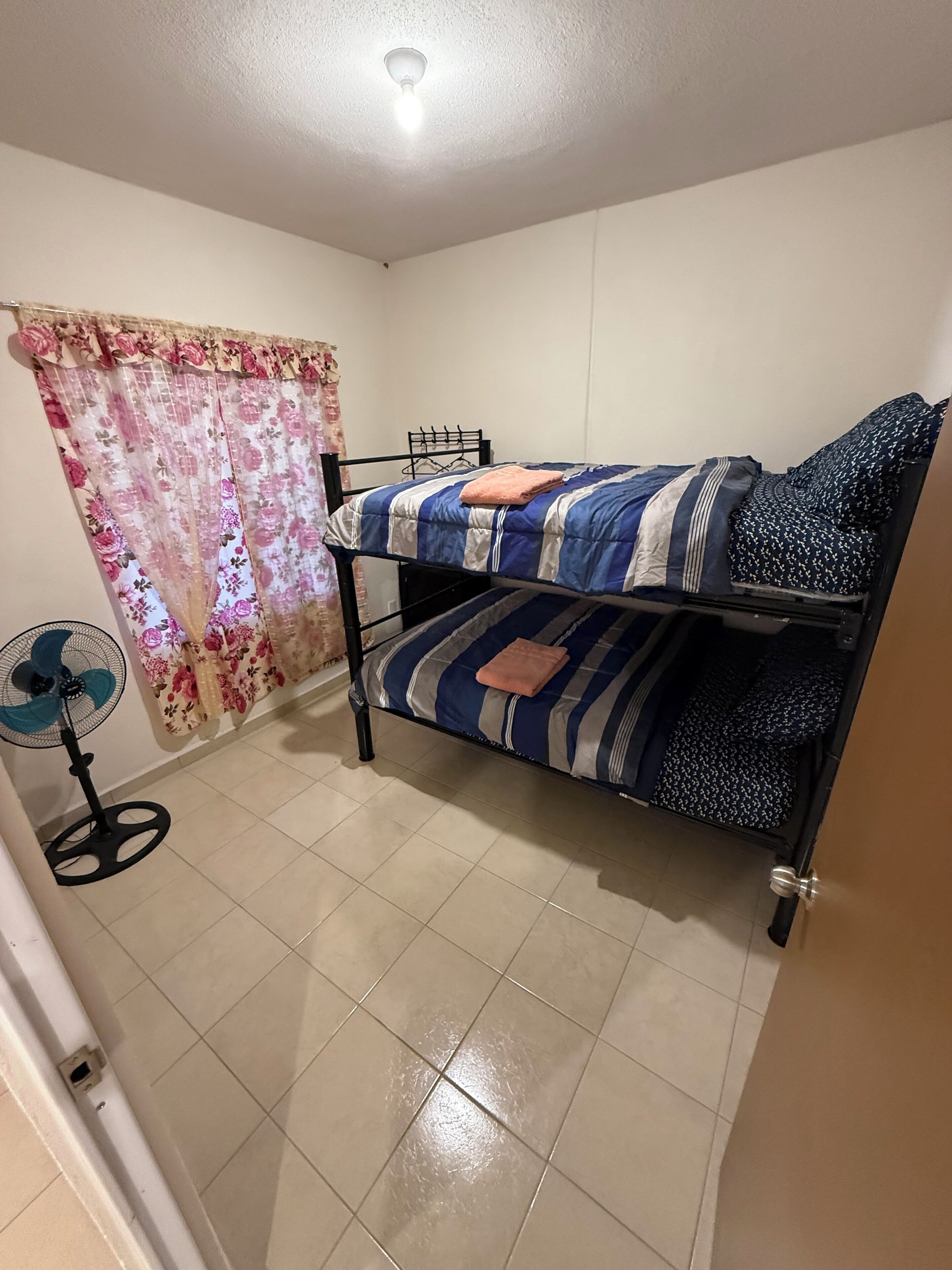 Una habitación pequeña y limpia con una cama individual, un ventilador de pie y una cortina estampada con motivos florales.