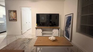 Una sala de estar moderna con una mesa de centro de madera, un mueble blanco para televisión y un amplio ventanal con persianas.