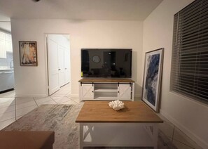 Una sala de estar moderna con una mesa de centro de madera, un mueble blanco para televisión y un amplio ventanal con persianas.