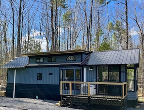 A small, single-story cabin with a metal roof, large windows, and a deck area. - Cozy 2-bedroom Tiny Home with sauna, A/C, boat ramp access to quiet lake (Casco)